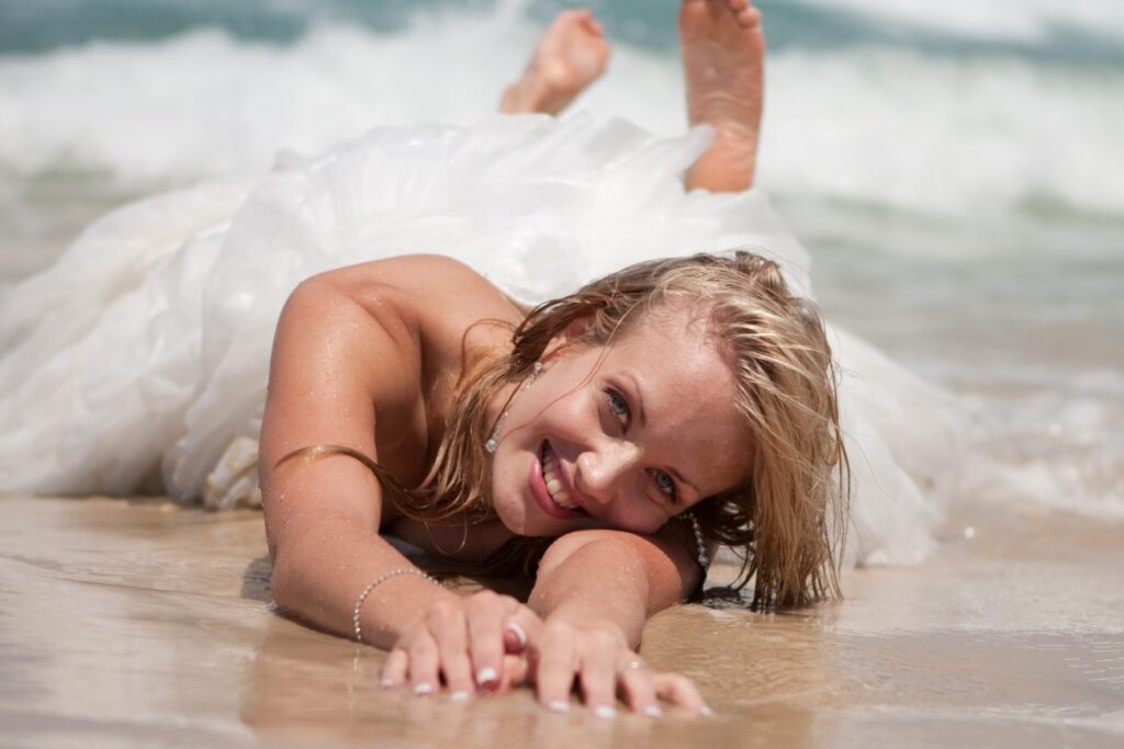 Woman in wedding dress laying on beach surf.