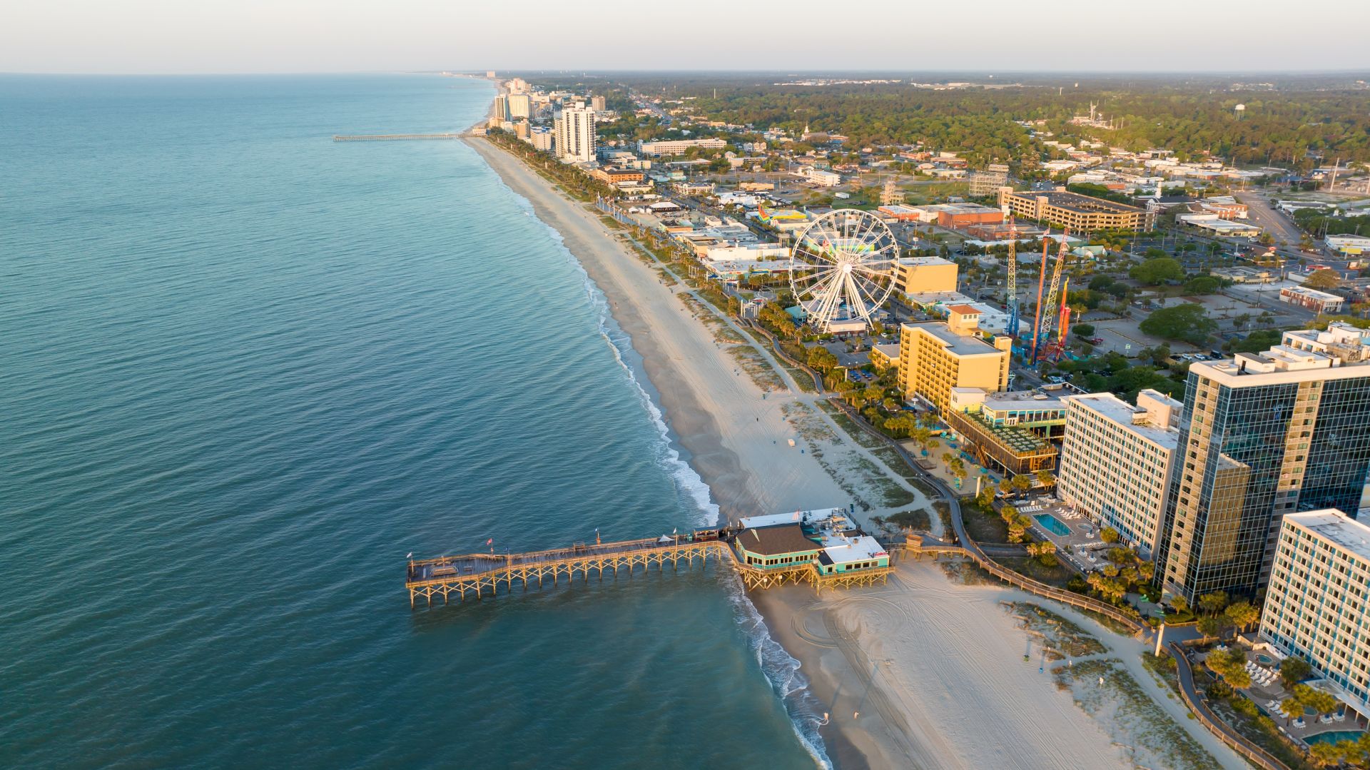 Aerial view of a sunny seaside city with a long white-sand beach, a pier, and a large Ferris wheel among modern buildings by the shore