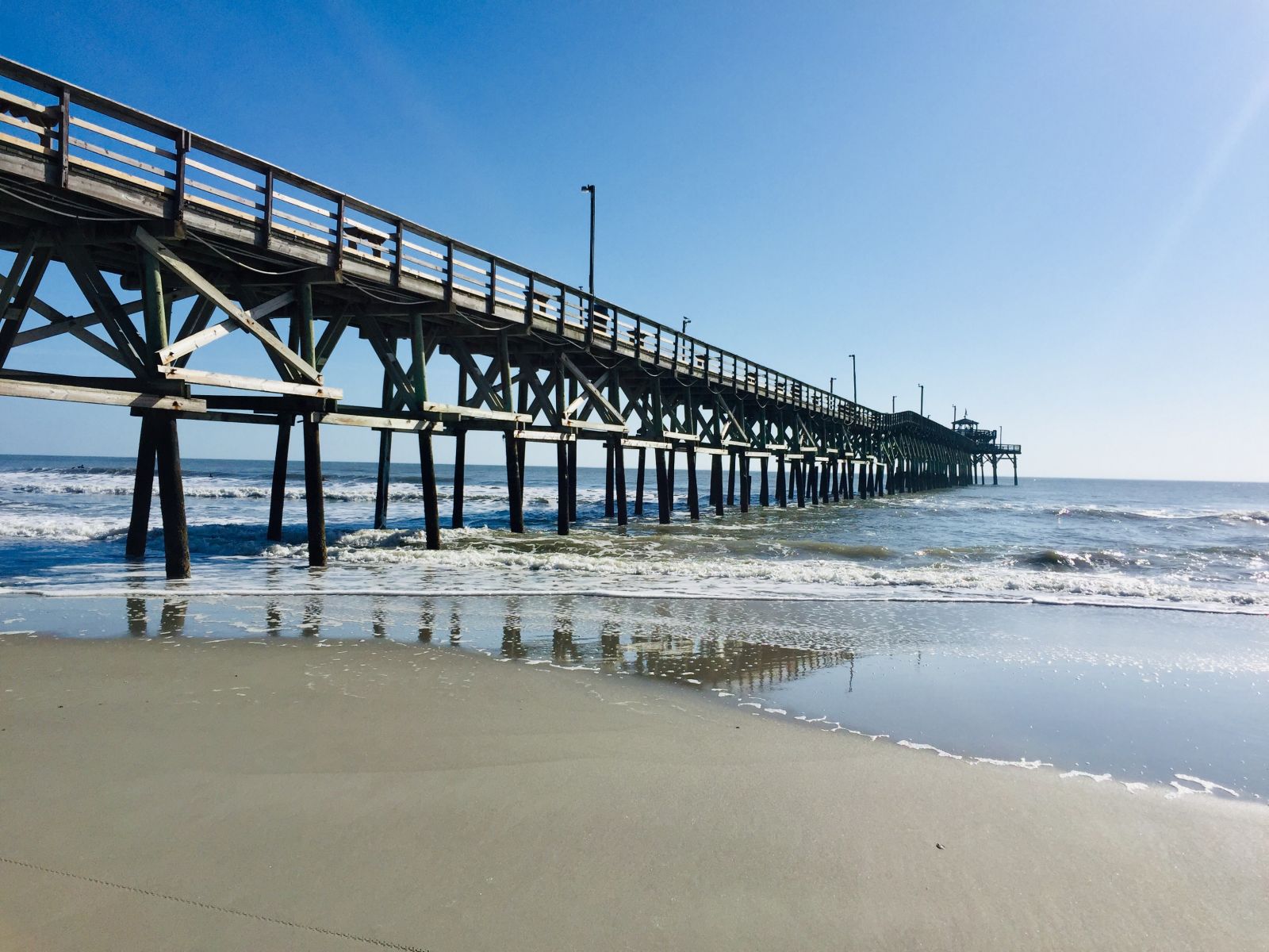 Long wooden pier extends from the beach into the blue ocean under a clear sky, with waves washing around its pilings.